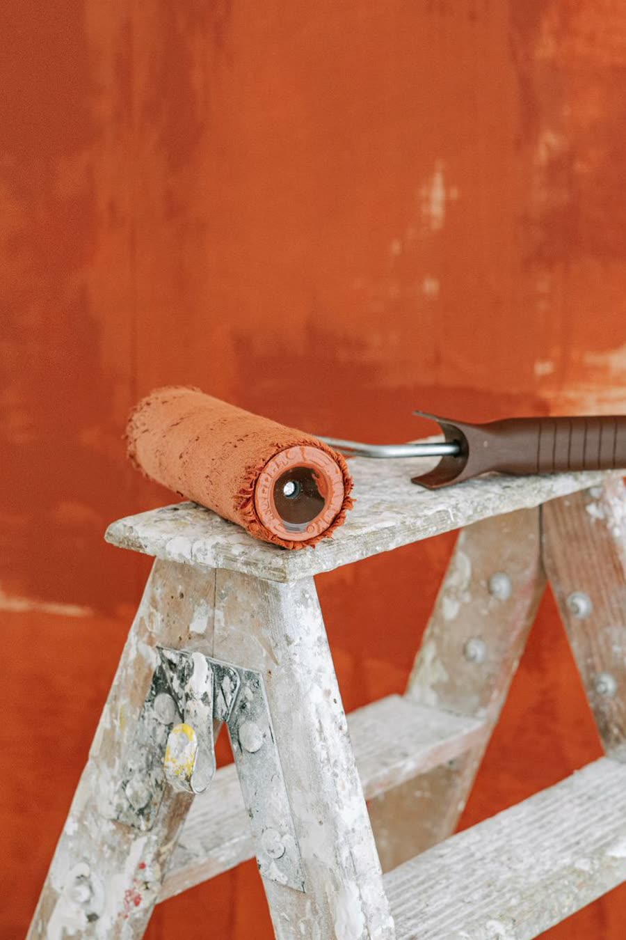 A paint roller resting on a spattered wooden stepladder against a half-painted terracotta wall